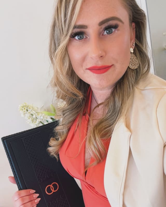 Pauline Hartmann lächelt in die Kamera und hält eine dunkle Trauungsmappe mit einem Blumenzweig.